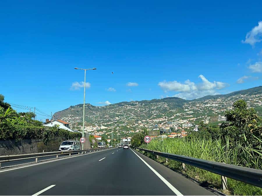 Driving on a sunny day along the VR1 highway in Madeira with a view of hillside homes.