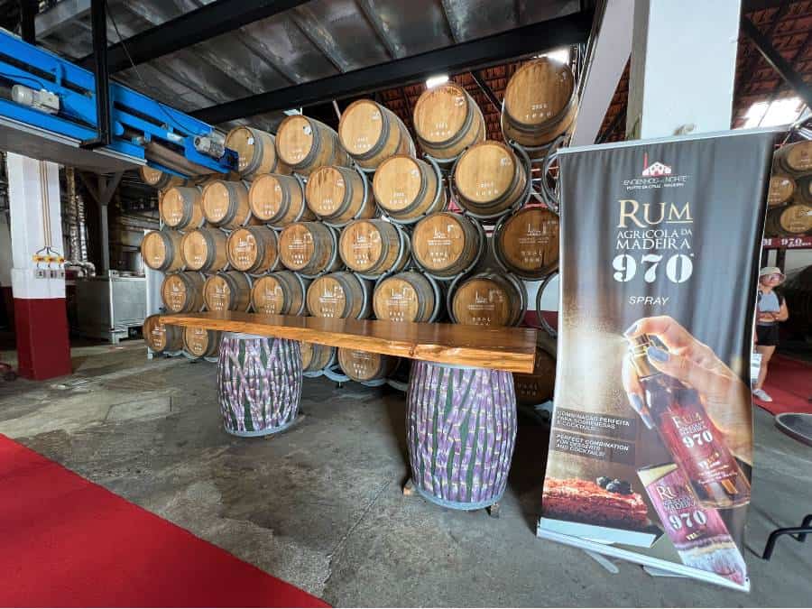 Tasting counter inside Engenhos do Norte distillery with a wall of rum barrels behind and promotional banner for Rum Agrícola da Madeira 970