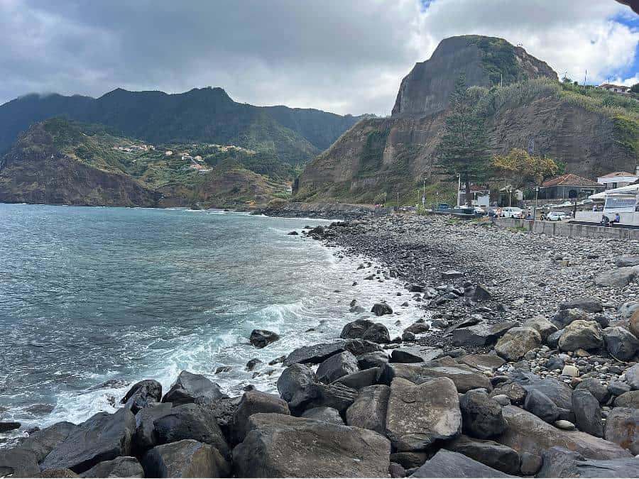 Rocky shoreline of Praia do Porto da Cruz with cliffs and houses in the background.