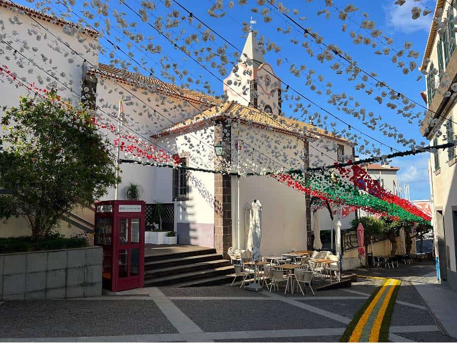 Outdoor café seating in a decorated town square in Ponta do Sol, Madeira, with a white church and red phone booth