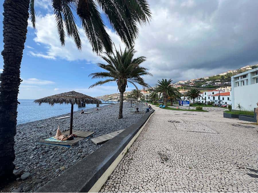 Palm-lined promenade and pebble beach in Santa Cruz, Madeira, with white houses and hills rising behind the town