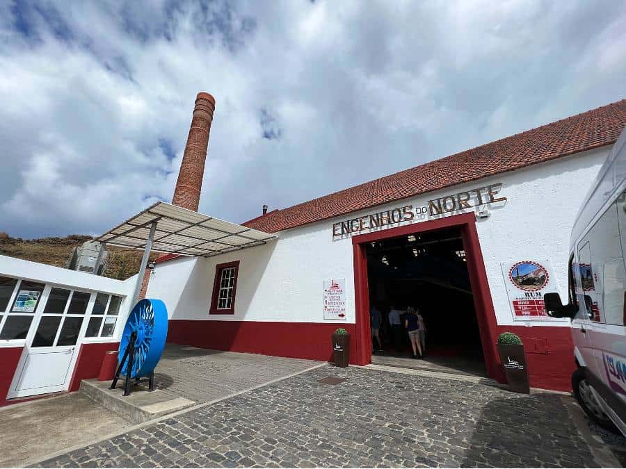 Exterior of Engenhos do Norte rum distillery in Porto da Cruz, Madeira, with tall brick chimney and visitors entering the building