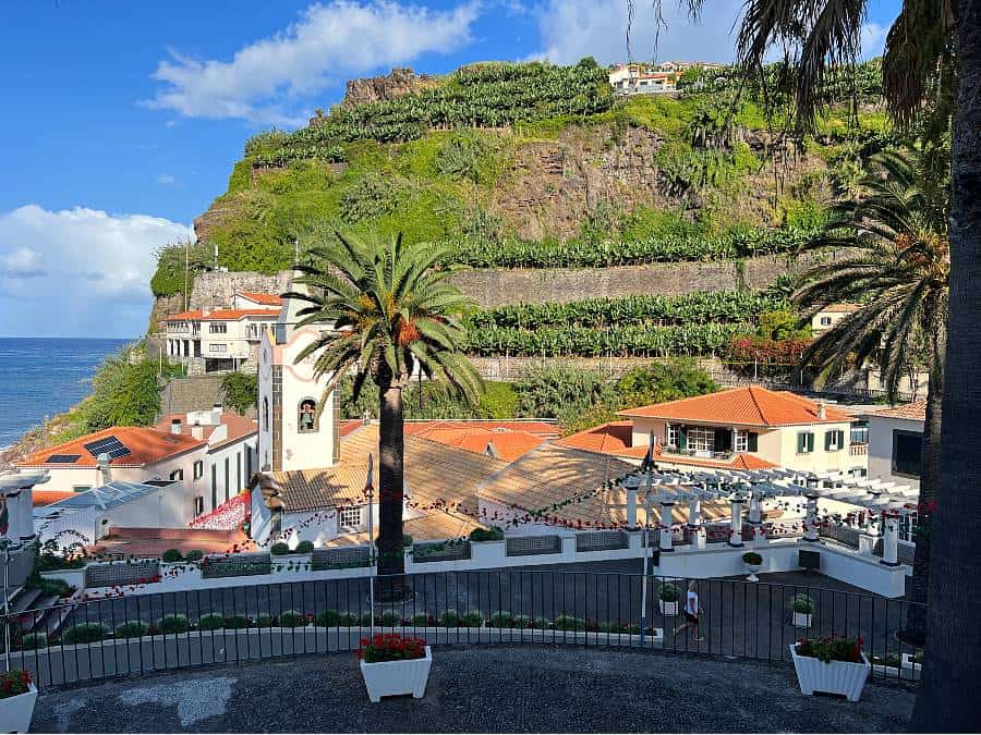 View over Ponta do Sol, Madeira, showing the white church of Nossa Senhora da Luz, red-roofed buildings, palm trees, and terraced banana fields on a green hillside.
