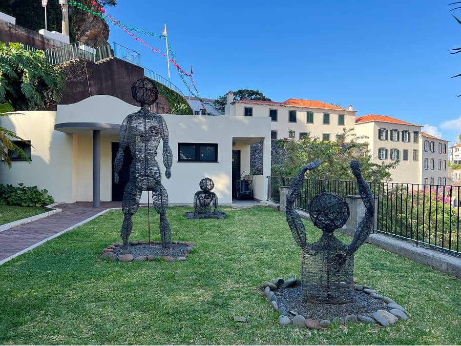 Three wire-frame human sculptures on grass in Ponta do Sol, part of a public art installation near the seafront.