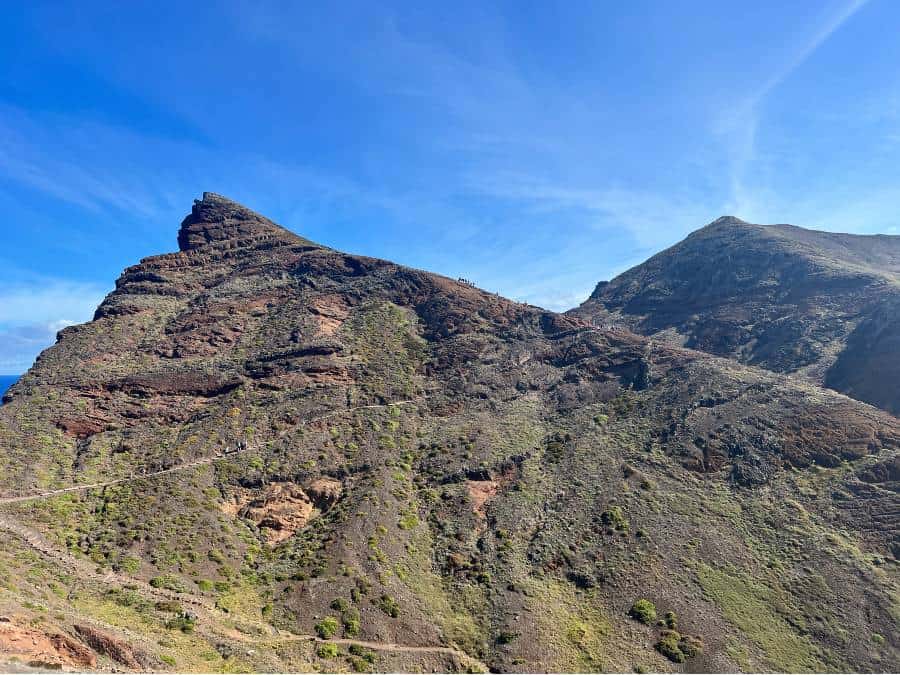 A rugged mountain peak with a winding hiking trail climbing up its slope under a clear blue sky on the Vereda da Ponta de São Lourenço trail in Madeira.
