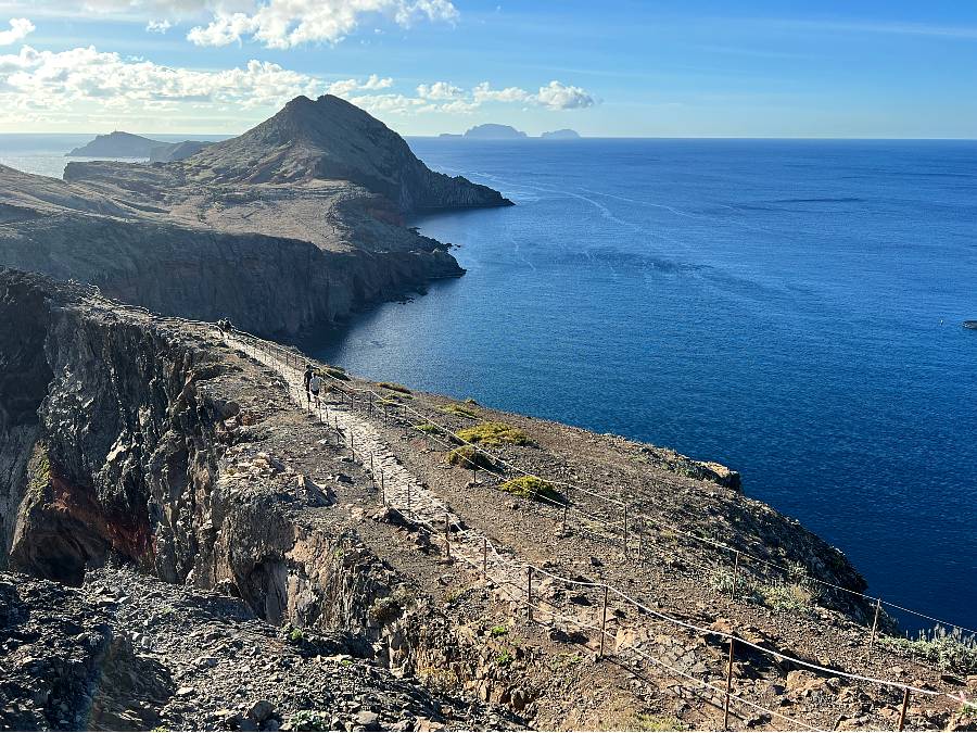 A rocky trail lined with safety cables winds along a dramatic cliffside with the deep blue Atlantic Ocean below and rugged peaks ahead on the São Lourenço peninsula.