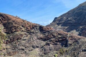 A long line of hikers makes their way up a rugged trail on the rocky hillside of the Vereda da Ponta de São Lourenço under a clear blue sky.