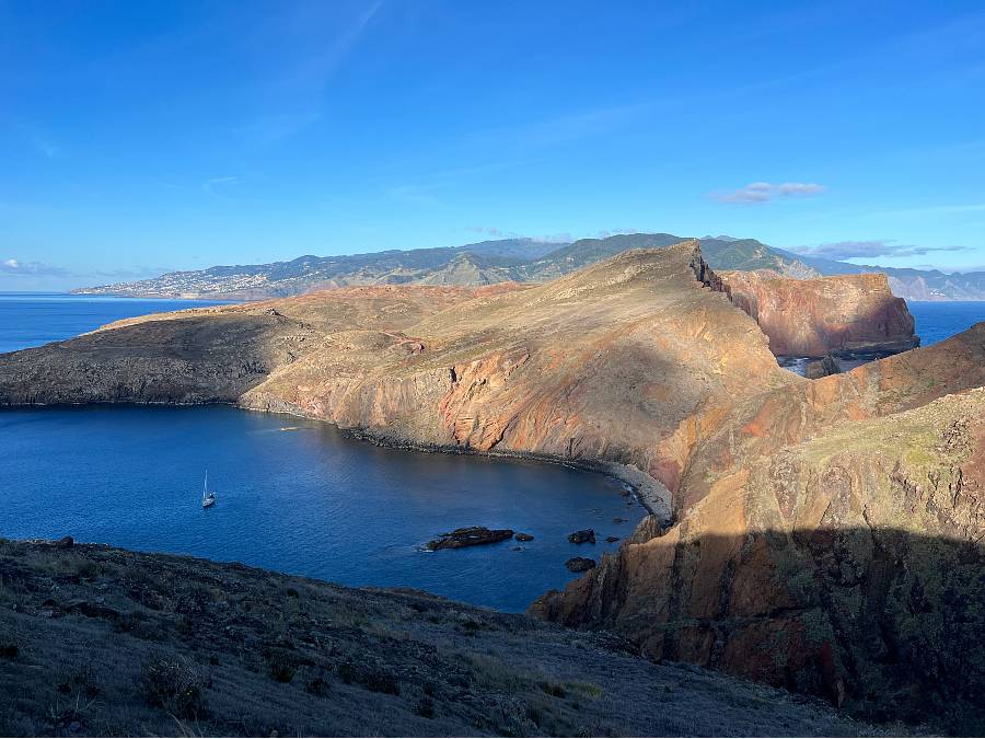 View of the São Lourenço peninsula with dramatic cliffs, calm bay waters, and a sailboat anchored near the shoreline, with Madeira’s mountainous interior in the background.